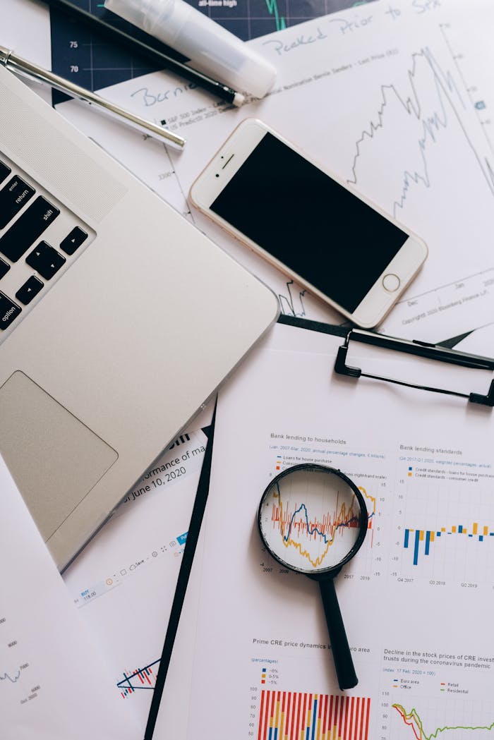 A detailed view of a workspace with laptop, smartphone, documents, and magnifying glass for financial analysis.